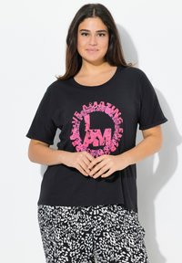 Černé triko s krátkým rukávem s růžovou květinovou grafikou s nápisem "JSEM ÚŽASNÝ" umístěným na hrudi; nošeno s vzorovanými kalhotami v černobílé barvě.