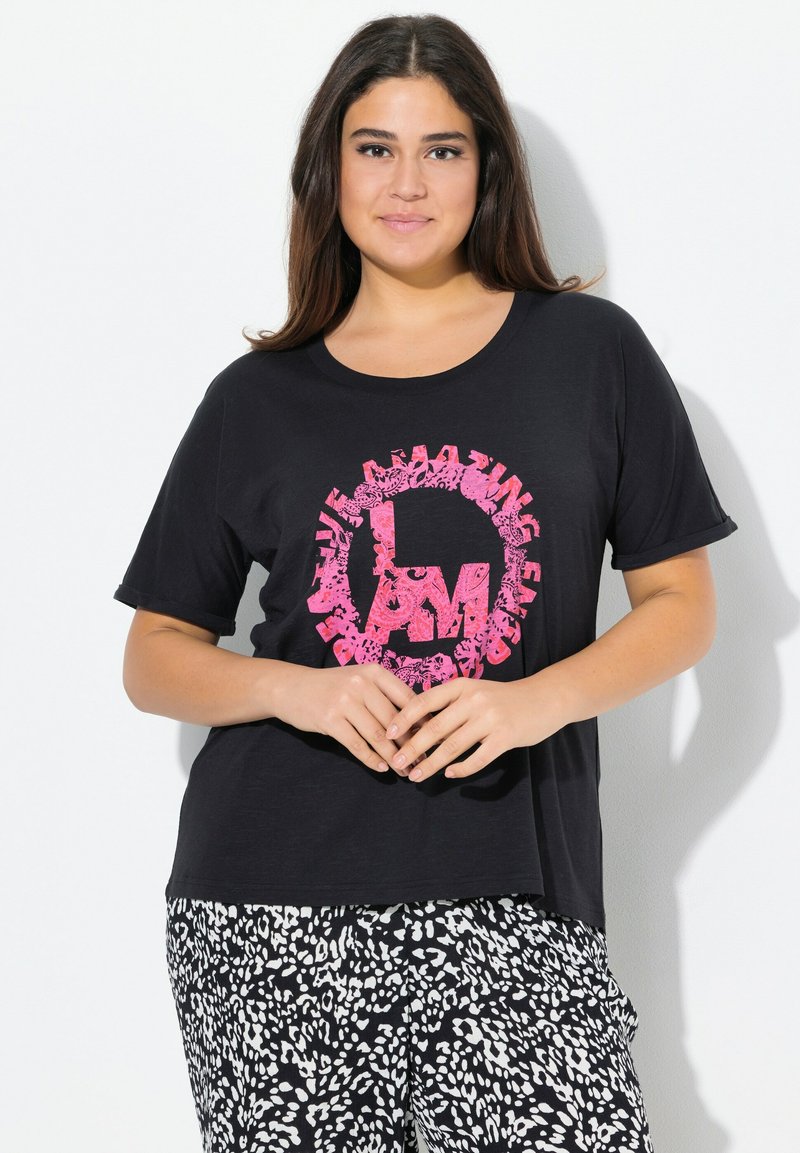 Černé triko s krátkým rukávem s růžovou květinovou grafikou s nápisem "JSEM ÚŽASNÝ" umístěným na hrudi; nošeno s vzorovanými kalhotami v černobílé barvě.