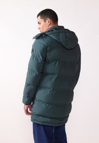 Giacca a vento verde con cappuccio, caratterizzata da un design testurizzato a trapunta, bottoni a scatto e un taglio lungo, visibile dalla schiena.