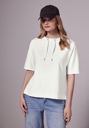 Felpa bianca a maniche corte con cappuccio regolabile e dettagli in metallo, abbinata a jeans blu chiaro e un cappellino da baseball nero.