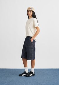 Cremefarbenes Kurzarm-T-Shirt, dunkelblaue Jeansshorts mit sichtbaren Nähten und Taschen, beigefarbene Kappe mit Logo, schwarze Turnschuhe, weiße Socken.