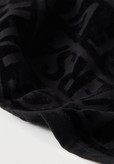Tissu noir avec des lettres en relief texturées formant un motif abstrait, présenté drapé sur une surface blanche.