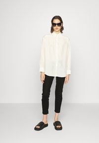 Chemise blanche à manches longues, pantalon noir ajusté, et sandales à glissière noires. Des lunettes de soleil complètent la tenue. Design simple et moderne.
