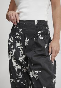 Czarne spodnie w kwiaty posiadają marszczony pas, lekki materiał oraz luźny krój z białymi wzorami kwiatów na całej powierzchni.