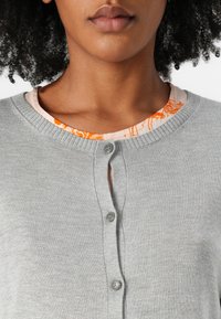 Šedý cardigan s kulatým výstřihem, zdobený zapínáním na knoflíky vpředu, přes světle oranžový vzorovaný top. Hladká textura, pletená tkanina.
