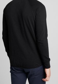 Svart långärmad tröja med rund halsringning, raglanärmar och en slät yta. Bakvy visar en något rundad nederkant.