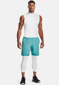 Weißes ärmelloses Oberteil, kombiniert mit türkisfarbenen Shorts und weißen Kompressionsleggings. Mit Under Armour-Logo auf Shorts und Oberteil. Schwarze Sportschuhe.