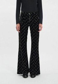 Pantalons évasés en velours noir avec un petit motif floral jaune, dotés d'une taille haute et de cinq boutons noirs pour la fermeture.