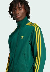 Zelená sportovní bunda se žlutými pruhy, vysokým límcem, zipem vpředu a kapsou na levé straně. Obsahuje adidas logo ve žluté barvě.