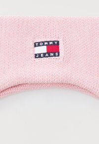 Pletena ružičasta kapa s preklopom. Ima pravokutnu oznaku s natpisom "TOMMY JEANS" i logotipom u mornarsko plavoj, bijeloj i crvenoj boji.