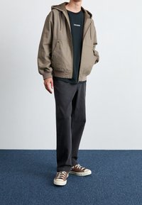 Khaki hooded jacket s ribbed cuffs, tmavé tričko s bílým textem, šedé kalhoty a hnědé tenisky s bílou podrážkou.