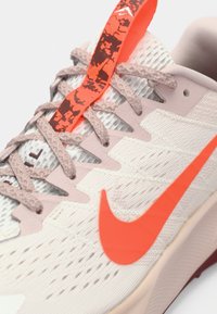 Gros plan d'une chaussure de course beige avec des lacets texturés, un logo Nike Swoosh orange vif et une languette à motif.
