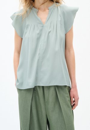 Vrouw draagt een lichtgroene blouse met korte mouwen en plooien, gecombineerd met loszittende olijfgroene broek.