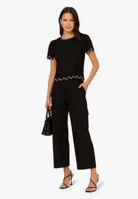 Černý krátký rukáv top s vlnitým lemem a perlovými detaily, v kombinaci s černými vysokými cargo kalhotami. Doplněno mini taškou a podpatky.