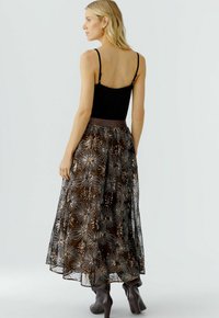 Maxi-Rock aus durchscheinendem braunem Stoff mit floralem, metallic glänzendem Stickerei. Figurnahes schwarzes Top mit verstellbaren Trägern und knielangen braunen Stiefeln.