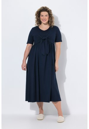 Smilende kvinde med krøllet hår iført en marineblå midi-kjole med sløjfe foran, korte ærmer og hvide slip-on sko, stående mod en hvid væg.