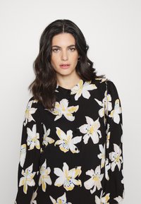 Blusa preta com grande padrão floral branco e amarelo, frente com botões e colarinho arredondado. Mangas longas, ligeiramente bufantes. Textura suave.