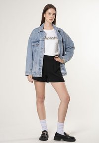 Lyseblå denim oversized jakke, hvid t-shirt med sort tekst, sorte plisserede shorts og sorte loafers med kædedetalje. Hvide sokker.