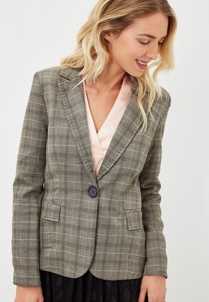 Mujer con un blazer de cuadros gris y un solo botón negro sobre una blusa rosa pálido, mirando hacia abajo y sonriendo levemente.