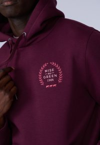 Hoodie bordeaux avec un logo rose brodé comprenant un design de laurier, le texte "MISE AU GREEN 1986" et trois petites vaches graphiques en bas.