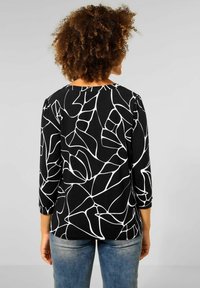 Svart topp med ett vitt abstrakt linjemönster, 3/4 ärmar och en avslappnad passform. Parat med blå jeans, som visar en lagerdesign.