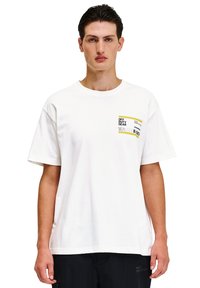 Hvid bomulds T-shirt med korte ærmer og rund hals. Har et rektangulært grafisk motiv i gul, sort og hvid på venstre brystområde.