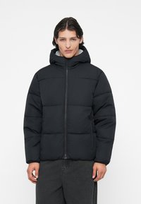 Jachetă neagră tip puffer cu glugă, având un fermuar frontal, textură matlasată și manșete elastice. Purtați-o peste o piesă de vestimentație gri, cu un design ușor.