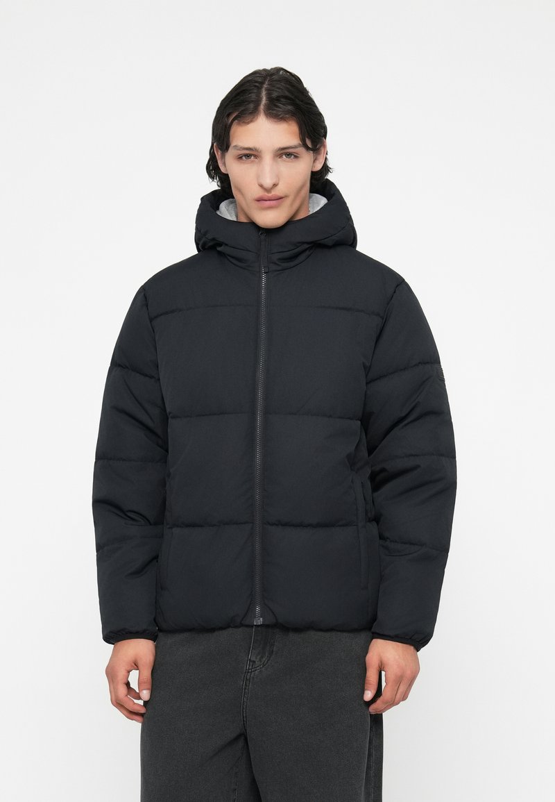 Jachetă neagră tip puffer cu glugă, având un fermuar frontal, textură matlasată și manșete elastice. Purtați-o peste o piesă de vestimentație gri, cu un design ușor.