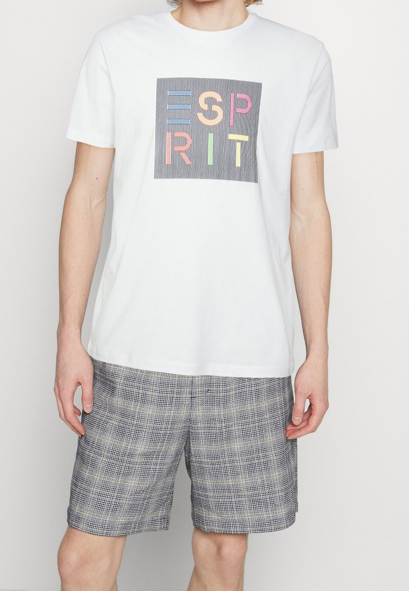 Camiseta blanca de algodón con un gráfico de letras coloridas sobre un fondo a rayas. Combinada con pantalones cortos de cuadros grises, de tela texturada.