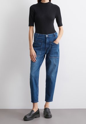 Vrouw draagt een zwarte top met korte mouwen, high-waisted blauwe jeans met grote voorzakken en zwarte instapschoenen, staand tegen een witte muur.
