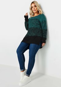 Swteter dzianinowy w kolorze niebiesko-zielonym i czarnym o teksturowanym wykończeniu, zestawiony z ciemnoniebieskimi dżinsami i białymi trampkami. Sweter ma luźny krój i dłuższy tył.