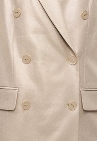 Μπεζ, διπλόκουμπος σακάκι με γυαλιστερό υφή υφάσματος. Διαθέτει χρυσά κουμπιά και δομημένο σχέδιο. Περιλαμβάνει πλαϊνές τσέπες.