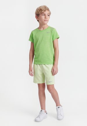 Blond chlapec stojí, nosí svetlozelené tričko a šortky Nike, biele tenisky Nike, pozerá sa doľava na bielom pozadí.