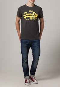 Camiseta gris con el logo amarillo de "SuperDry", jeans azules y zapatillas rojas con suelas blancas. Diseño sencillo, estilo casual.