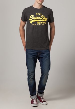 Camiseta gris con el logo amarillo de "SuperDry", jeans azules y zapatillas rojas con suelas blancas. Diseño sencillo, estilo casual.