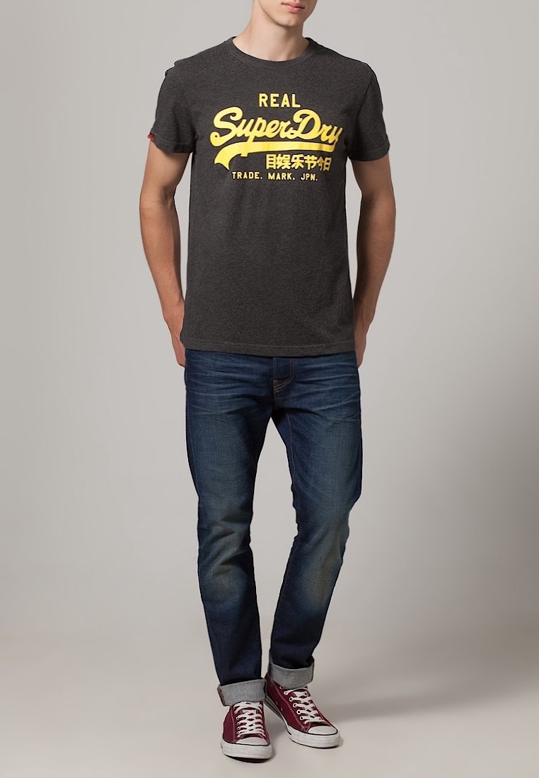 Camiseta gris con el logo amarillo de "SuperDry", jeans azules y zapatillas rojas con suelas blancas. Diseño sencillo, estilo casual.