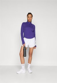 Lilla langermet treningstopp med halv glidelås, kombinert med hvite shorts, hvite joggesko og holder en svart vannflaske.