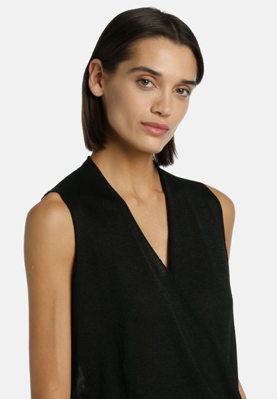 Femme aux cheveux bruns courts portant un haut noir sans manches à col en V, légèrement tournée vers la droite sur un fond blanc uni.