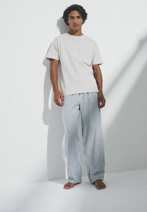 Noor mees seisab paljajalu valge seina taustal, kandes lihtsat valget t-särki ja lahtisi sinivalgekirju pükse, vasakul käsivarrel tätoveering.