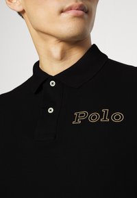 Svart polo skjorta med texturerad tyg, två knappar och broderad "Polo"-logotyp i guld på bröstet. Skräddarsydd krage synlig.
