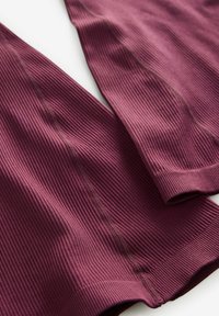 Ribbede maroon stof, der viser to overlappende sektioner med sømløse kanter og tekstureret overflade, der fremhæver tonale variationer.