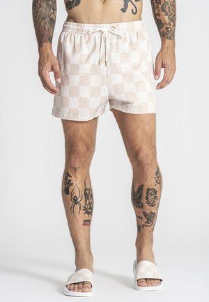 Mand iført beige og hvide ternede badebukser og matchende klipklapper, der viser forskellige sorte tatoveringer på ben og arme.