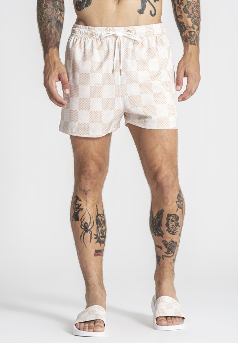 Mies, jolla on beigevalkoisetruudulliset uimashortsit ja niihin sopivat liukusandaalit, esittelee erilaisia mustia tatuointeja jaloissa ja käsivarsissa.