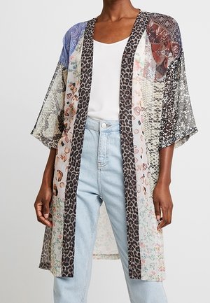 Person iført en gennemsigtig, knælængde patchwork kimono-stil jakke med blandede dyre- og blomstermønstre over en hvid top og lyseblå jeans.