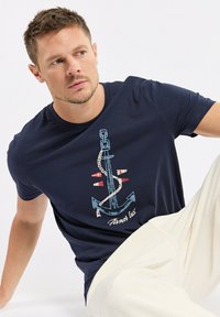 T-shirt en coton bleu marine avec un graphisme d'ancre, de la corde et des accents rouges. Style col rond et manches courtes avec une coupe décontractée.