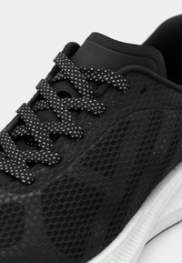 Κοντινή λήψη ενός μαύρου αθλητικού παπουτσιού με υφασμένο πάνω μέρος, μαύρα κορδόνια με λευκές τελείες και λευκή αντικραδασμική σόλα.