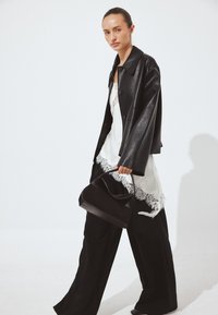 Nainen kävelee musta nahkatakki yllään valkoisen pitsireunaisen topin ja leveiden mustien housujen kanssa, kantaen mustaa käsilaukkua kädessään.