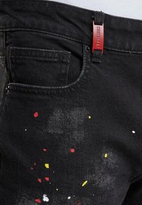 Czarne dżinsy z przodu z kieszenią, ozdobione wzorem w plamy czerwonej, żółtej i białej farby. Czerwona metka z logo przyszyta do pasa.
