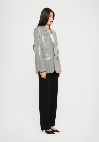 Donna in piedi che indossa un blazer grigio a quadri con paillettes sottili, un top bianco, pantaloni larghi neri e tacchi a punta neri contro uno sfondo semplice.