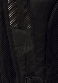Detail van een zwarte rugzak met gaasstof en glad textiel, voorzien van een band en logo label. Accentuelementen omvatten een gesp en stiksels.